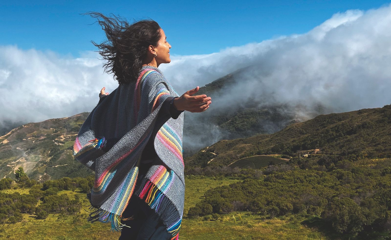 Lourdes Vallenzuela Allende, de pie en un cerro verde con los brazos abiertos, entre nubes bajas y montanas.